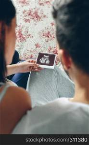 Couple looking at ultrasound of their baby sitting on the bed. Selective background focus on ultrasound. Couple looking at ultrasound of their baby