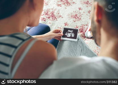 Couple looking at ultrasound of their baby sitting on the bed. Selective background focus on ultrasound. Couple looking at ultrasound of their baby