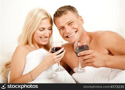 Couple lying in bed together sharing wine and smiling looking at camera