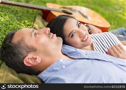 Couple lying on grass with acoustic guitar looking at camera smiling
