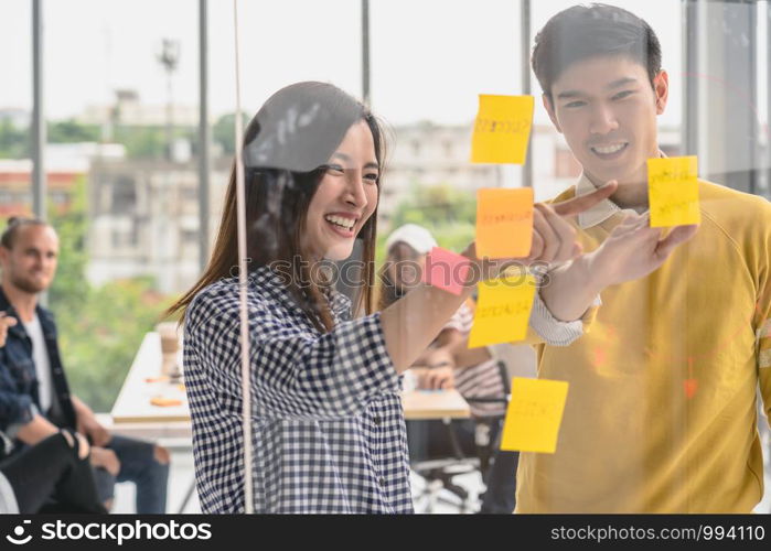 Couple of creative colleague writing and pointing the postit on the millor board when presenting the ideas over Group Of Asian Business people with casual suit in the modern workplace