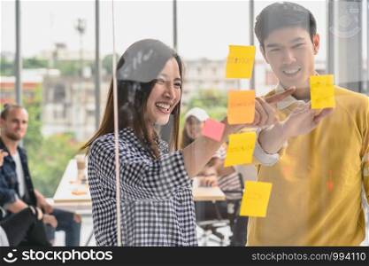 Couple of creative colleague writing and pointing the postit on the millor board when presenting the ideas over Group Of Asian Business people with casual suit in the modern workplace
