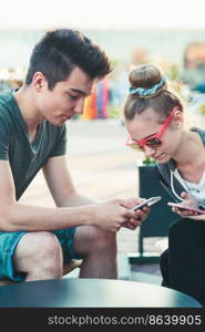 Couple of friends, teenage girl and boy,  having fun using smartphones sitting in center of town, spending time together