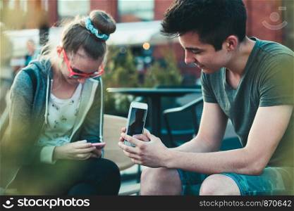 Couple of friends, teenage girl and boy, having fun using smartphones sitting in center of town, spending time together