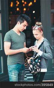 Couple of friends, teenage girl and boy, using smartphones, talking together, standing on street in center of town, spending time together
