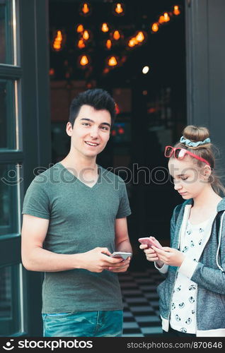 Couple of friends, teenage girl and boy, using smartphones, talking together, standing on street in center of town, spending time together