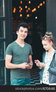 Couple of friends, teenage girl and boy, using smartphones, talking together, standing on street in center of town, spending time together