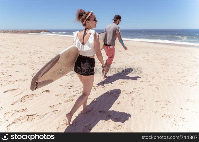 Couple of surfers with surfboards walking at a sandy beach.