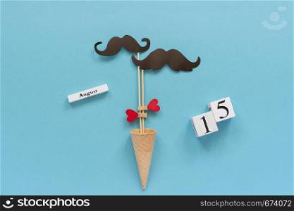 Couple paper mustache props fastened heart in ice cream cone and calendar 15 August on blue background. Concept Homosexuality gay love, International Gay Day.. Couple paper mustache props fastened heart in ice cream cone and calendar 15 August on blue background. Concept Homosexuality gay love, International Gay Day