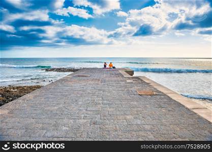 Couple people sitting on edge of stone pier looking at sea landscape