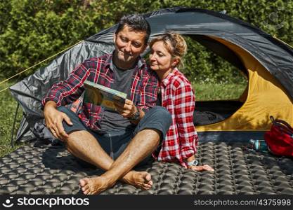 Couple planning next trip while sitting with map by tent. People relaxing at camping during summer vacation. Actively spending vacations outdoors close to nature. Concept of camp life