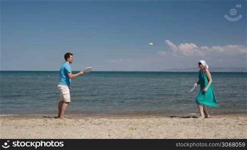 Couple playing bat and ball tennis game at the beach on the golden sand at the edge of a calm ocean as they enjoy their summer vacation