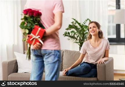couple, relationships and people concept - happy woman looking at man with flowers and gift at home. woman looking at man with flowers and gift at home