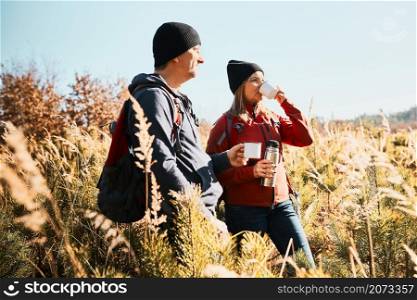 Couple relaxing and enjoying the coffee during vacation trip. People standing on trail drinking coffee. Friends with backpacks hiking through tall grass along path in mountains. Active leisure time close to nature