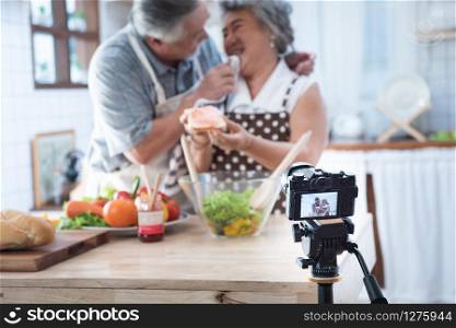 Couple senior Asian elder happy living in home kitchen. Grandfather wiping grandmother mouth after eating bread with jam vlog vdo for social blogger. Focus on camera. Modern lifestyle & relationship.