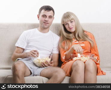 Couple sits on the couch. Each lap Cup of popcorn. Couple watching TV, eating popcorn and smiling.