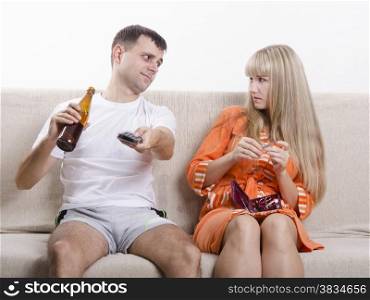 Couple sits on the couch. He is watching the TV, switch the channels with the remote, drinking beer, and grins, looking at her, she handles nails nail file and looked at him reproachfully