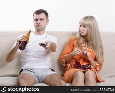 Couple sits on the couch. He is watching the TV, switch the channels with the remote, drinking beer, and grins, looking at her, she handles nails nail file and looked at him reproachfully