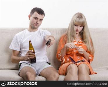 Couple sits on the couch. He watches TV, remote switch channels, drink beer, it handles nails nail file.