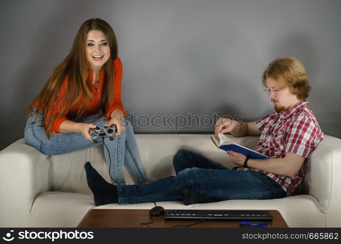 Couple sitting on couch spending free time. Woman playing video games and man reading book.. Woman playing games man reading book