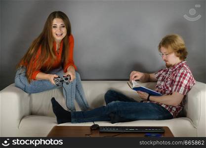 Couple sitting on couch spending free time. Woman playing video games and man reading book.. Woman playing games man reading book