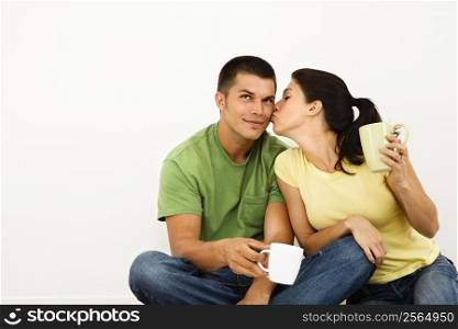 Couple sitting on floor drinking coffee with woman leaning over kissing man on cheek.