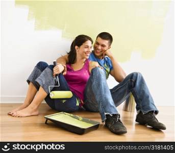 Couple sitting on floor smiling in front of partially painted wall in home.