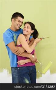 Couple smiling and embracing in front of partially painted interior wall holding paintbrush and roller.