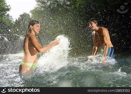 Couple splashing water at each other in forest river, side view