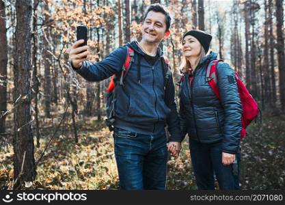 Couple taking selfie photo while vacation trip. Hikers with backpacks walking on path in forest on sunny day. Active leisure time close to nature