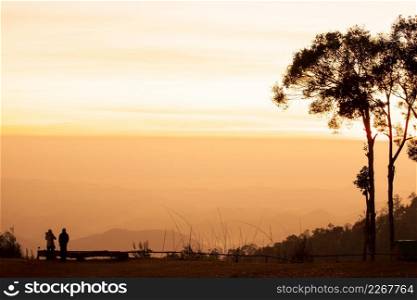 Couple tourists standing on the mountains and look at view in the morning, colorful scenic landscape, golden light shines down around the tropical forest. Winter season. Warm tone. Traveler, Journey.