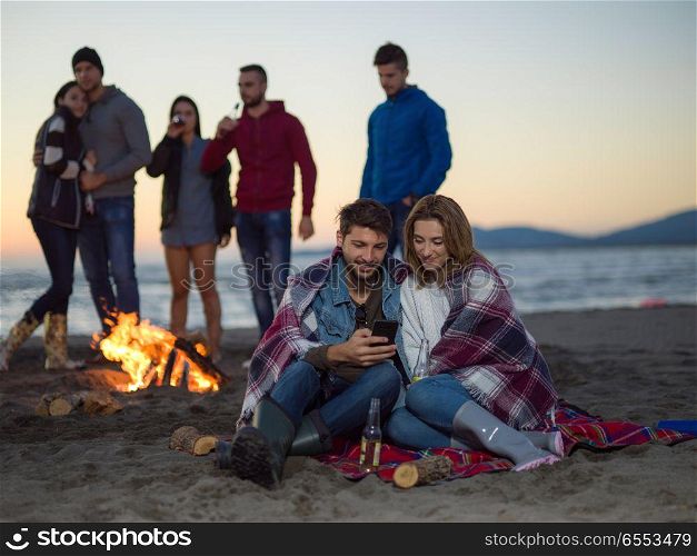 Couple using cell phone during autumn beach party with friends drinking beer and having fun. Couple enjoying bonfire with friends on beach