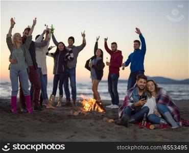 Couple using cell phone during autumn beach party with friends drinking beer and having fun. Couple enjoying bonfire with friends on beach