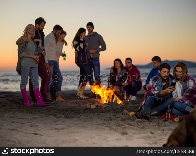 Couple using cell phone during beach party with friends drinking beer and having fun. Couple enjoying bonfire with friends on beach