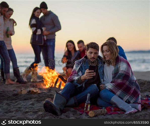 Couple using cell phone during beach party with friends drinking beer and having fun. Couple enjoying bonfire with friends on beach