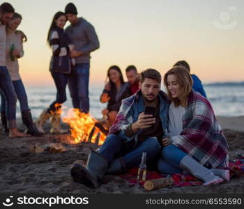 Couple using cell phone during beach party with friends drinking beer and having fun. Couple enjoying bonfire with friends on beach