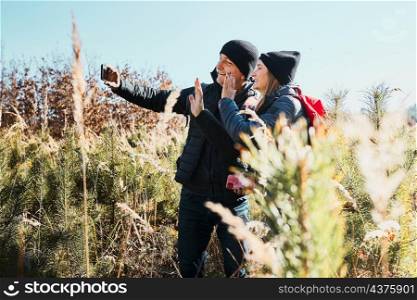 Couple waving during video call sending greetings from vacation trip. Hikers with backpacks walking through tall grass along path in meadow on sunny day. Active leisure time close to nature