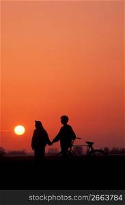 Couple with bicycle holding hands silhouetted against sunset sky
