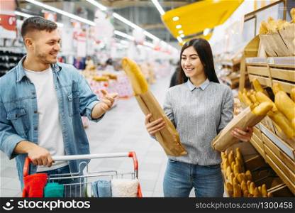 Couple with cart choosing fresh loaf in a supermarket, family shopping. Customers in shop, buyers in market. Couple with cart choosing loaf in a supermarket