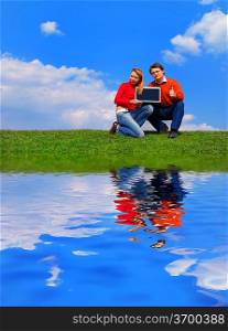 Couple with notebook sitting on grass against sky with reflection on water