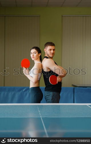 Couple with rackets poses at the ping pong table with net indoors. Man and woman in sportswear, table-tennis training game in gym