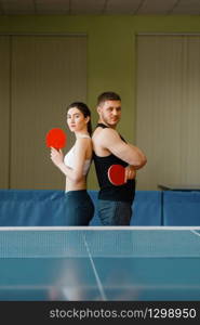 Couple with rackets poses at the ping pong table with net indoors. Man and woman in sportswear, table-tennis training game in gym