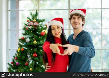 Couple young man and woman use hands to make symbol of heart and they stay in room with decoration of Christmas festival with happiness.