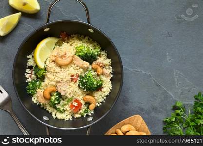 Couscous dish with chicken, broccoli, tomato, cashew nuts. Lemon, cashew nuts and parsley on the side, photographed overhead on slate with natural light.