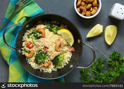 Couscous dish with chicken, broccoli, tomato, cashew nuts. Lemon, cashew nuts, salt, and parsley on the side, photographed overhead on slate with natural light.