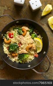 Couscous dish with chicken, broccoli, tomato, cashew nuts. Lemon, salt and pepper shakers on the side, photographed overhead on slate with natural light.