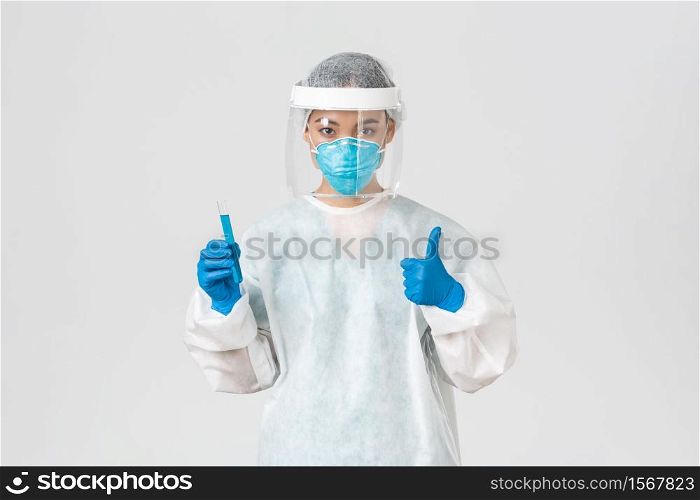 Covid-19, coronavirus disease, healthcare workers concept. Confident serious asian female doctor, tech lab employee in personal protective equipment show thumb-up and hold test-tube with vaccine.. Covid-19, coronavirus disease, healthcare workers concept. Confident serious asian female doctor, tech lab employee in personal protective equipment show thumb-up and hold test-tube with vaccine