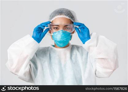 Covid-19, coronavirus disease, healthcare workers concept. Serious-looking asian female doctor in personal protective equipment, put on glass before entering lab, white background.. Covid-19, coronavirus disease, healthcare workers concept. Serious-looking asian female doctor in personal protective equipment, put on glass before entering lab, white background