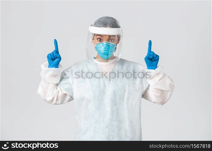 Covid-19, coronavirus disease, healthcare workers concept. Surprised and intrigued asian female doctor in respirator and personal protective equipment, pointing fingers up at advertisement.. Covid-19, coronavirus disease, healthcare workers concept. Surprised and intrigued asian female doctor in respirator and personal protective equipment, pointing fingers up at advertisement