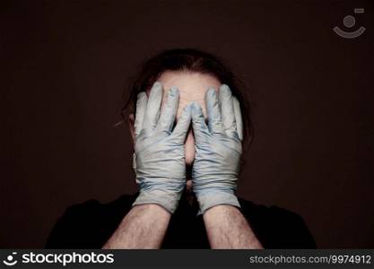 Covid-19 / Coronavirus. Human Portrait of a disease. Man with the head in the hands wearing protective blue sterile latex gloves